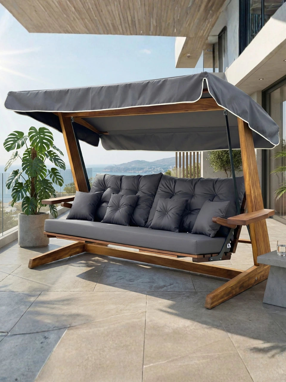 Leagăn de grădină Luna Premium din lemn, cu pernă și umbrelă gri antracit, pe terasă, cu decor format din plante ornamentale verzi si peisaj montan în plan îndepărtat.