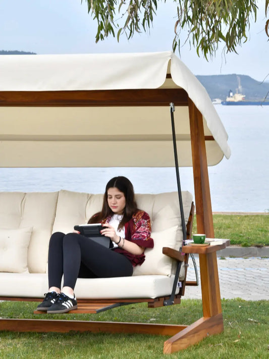 Persoană așezată într-un leagăn de grădină Luna Premium din lemn, cu perne și umbrelă bej,  într-un peisaj cu lac și munți