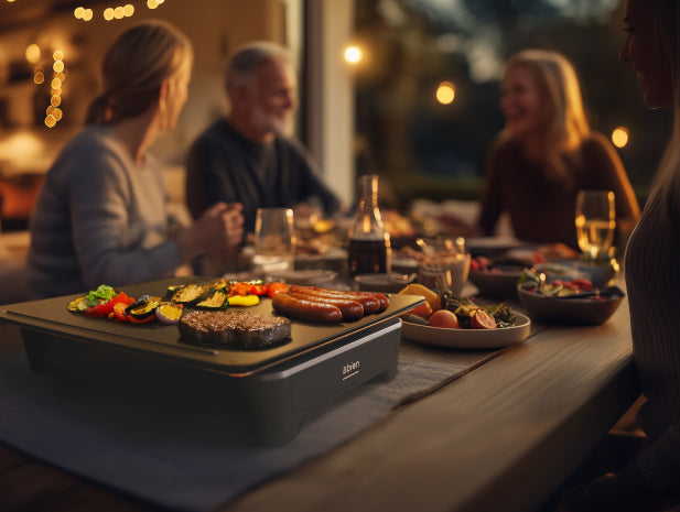 Oameni adunati în jurul unei mese cu mâncare și băuturi, pe care se afla o plita electrica portabila